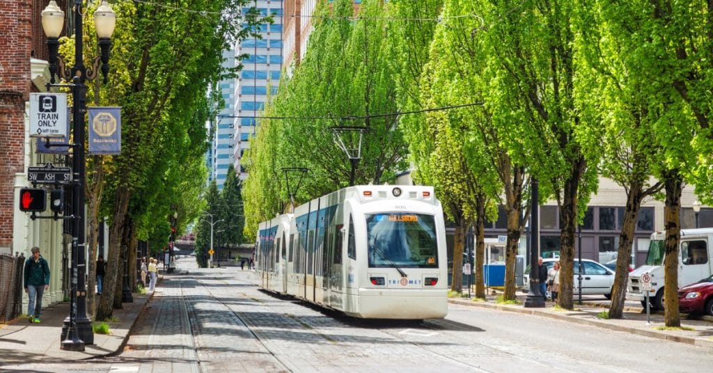 A light rail train moving beside a road with mature green trees, tall buildings, parked cars, and people on the sidewalks.
