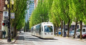 A light rail train moving beside a road with mature green trees, tall buildings, parked cars, and people on the sidewalks.