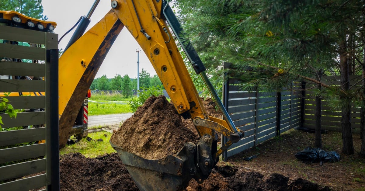 A backhoe digs into soil near established evergreen trees during excavation work on a residential property.